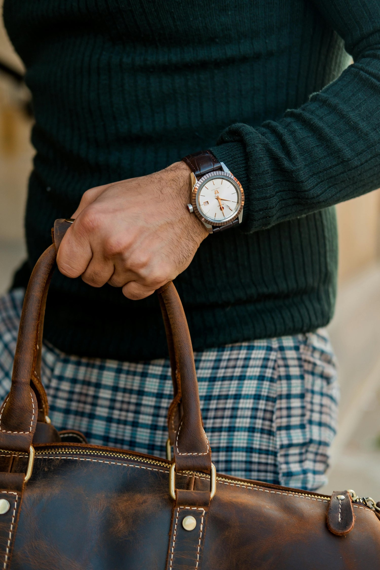 Elegante Vistora Seiko-Mod Herrenuhr mit weißem Zifferblatt, goldenen Akzenten und braunem Lederarmband. Perfekt für Business- und Casual-Looks. Getragen von einem Mann mit modisch modernem Outfit. Zu sehen ist ein Auschnitt, indem er die Seiko-Mod, eine Ledertasche, einen grünen Strickpullover und eine Karierte Hose trägt.
